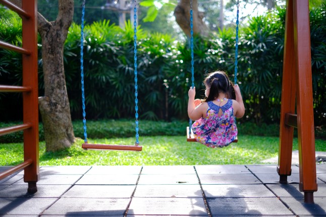 The back view of a lonely young Asian girl playing on a swing by herself in an empty playground on a sunny day. ; Shutterstock ID 1892000131; purchase_order: -; job: -; client: -; other: -