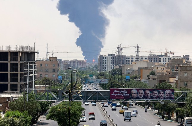 Portraits of Iranian military generals and nuclear scientists, killed in Israel's June 13 attack are displayed above a road, as a plume of heavy smoke and fire rise from an oil refinery in southern Tehran, after it was hit in an overnight Israeli strike, on June 15, 2025. Iran fired a fresh barrage of missiles at Israel, state television announced early on June 15, as the rivals exchanged fire for a third day. (Photo by ATTA KENARE / AFP) (Photo by ATTA KENARE/AFP via Getty Images)