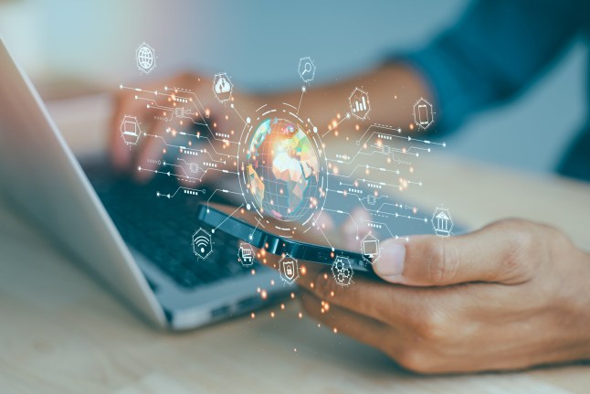 A person is using a laptop and a cell phone, both of which are connected to the internet. The laptop screen shows a globe with various icons