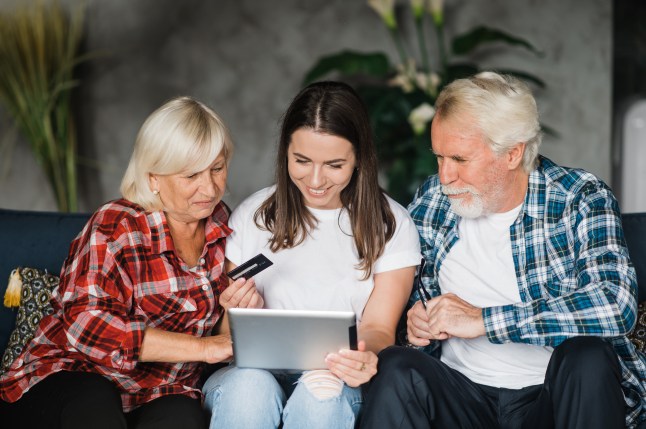 A young woman sat on a sofa looking at a tablet and holding a bank card with her parents sitting either side.
