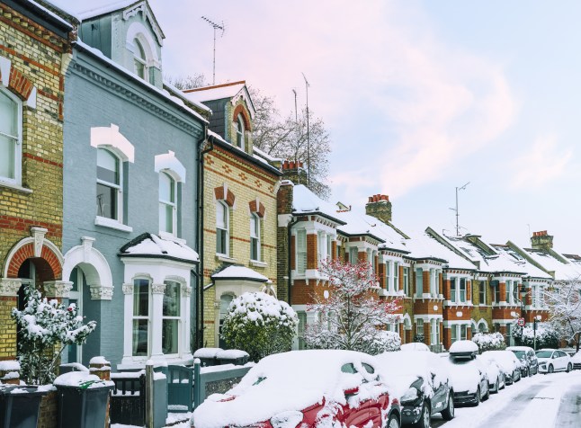 A road with a row of terraced houses and cars parked along it covered in snow under a blue sky.