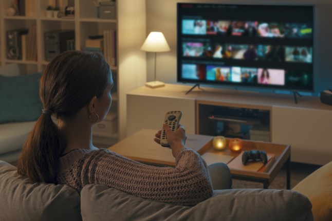 Woman relaxing on the couch, she is using the remote control and choosing a TV show or movie on the television menu; Shutterstock ID 2126878775; purchase_order: -; job: -; client: -; other: -