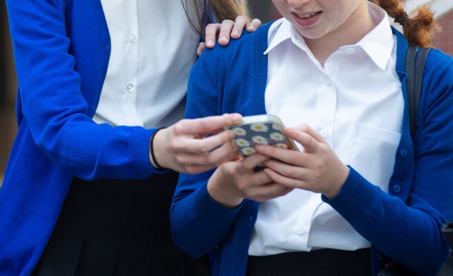 A three-quarters shot of a group of female teenage students in their school. They are smiling and talking to each looking down at a mobile phone. This school is based in the North East of England.