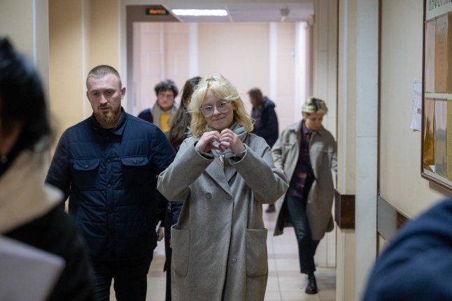 A young woman with bleach blonde hair wearing glasses and a long grey coat walks through a corridor surrounded by police