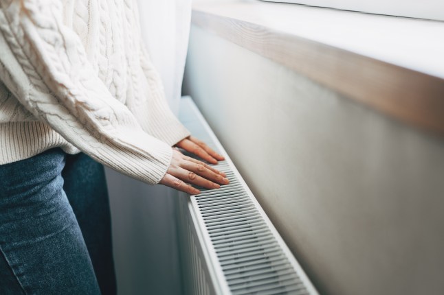 A woman wearing a white cable knit jumper and jeans placing herhands on a white radiator.