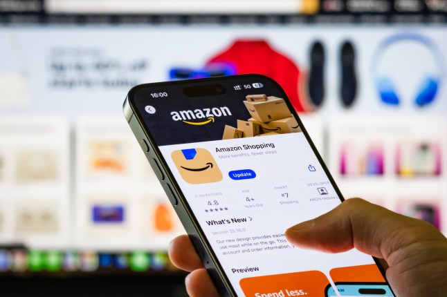 CHONGQING, CHINA - AUGUST 22: In this photo illustration, a person holds a smartphone showing the Amazon Shopping app page in the Apple App Store, with a computer screen displaying the Amazon website in the background on August 22, 2025 in Chongqing, China. (Photo illustration by Cheng Xin/Getty Images)