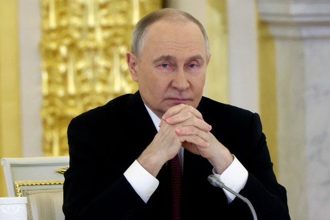 Putin Attends Council On National Projects. The background is gold and white. Putin wears a black jacket and maroon tie, clasps his hands together and looks directly at the camera.