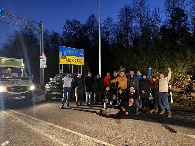 A host of volunteers for Driving Ukraine take a photo in front of the Ukraine border sign.