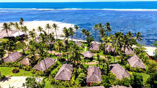 A beautiful thatched-roof bungalow, a traditional Fijian bure, sits on the edge of a white sand beach. The ultimate tropical island escape and luxury accommodation