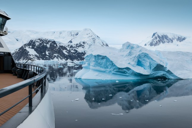 Scenic view of Neko Harbor in Antarctica with calm water reflecting snowy mountains and a drifting iceberg, photographed from an expedition cruise ship with railing visible.