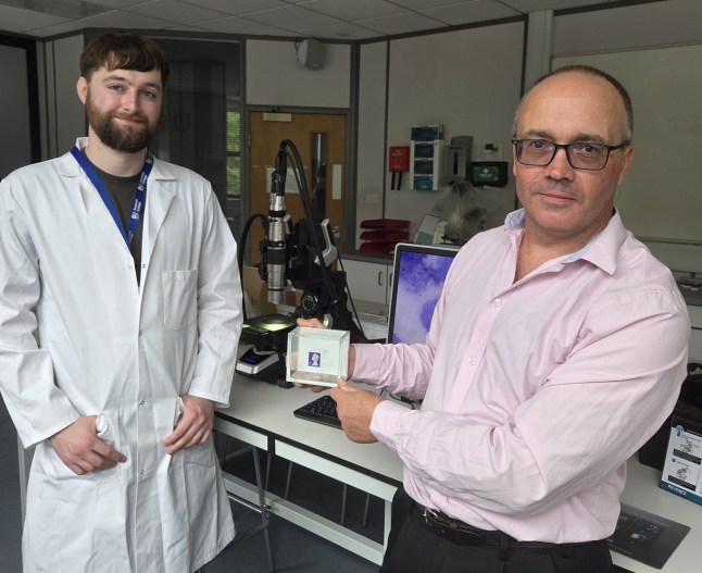 Man in a lab coat standing with a man wearing glasses and a pink shirt holding his micro sculpture creation set in a glass box