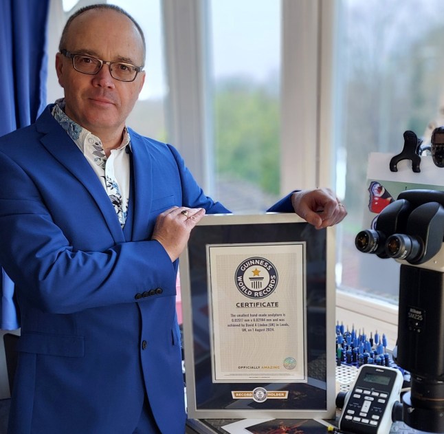 A man dressed in a royal blue blazer holding a Guinness World Record certificate for world's smallest handmade sculpture